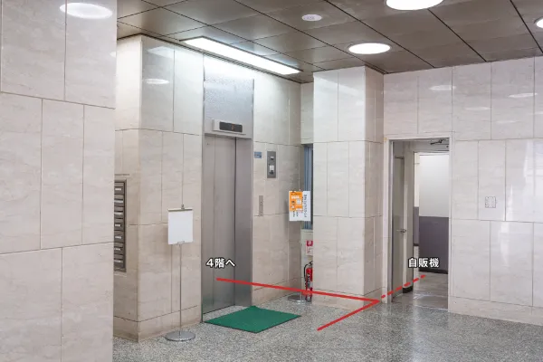 TOビル1階エントランス内部。中央にエレベーター、右奥に自販機の通路があり、4階への動線が赤い線で示されている
